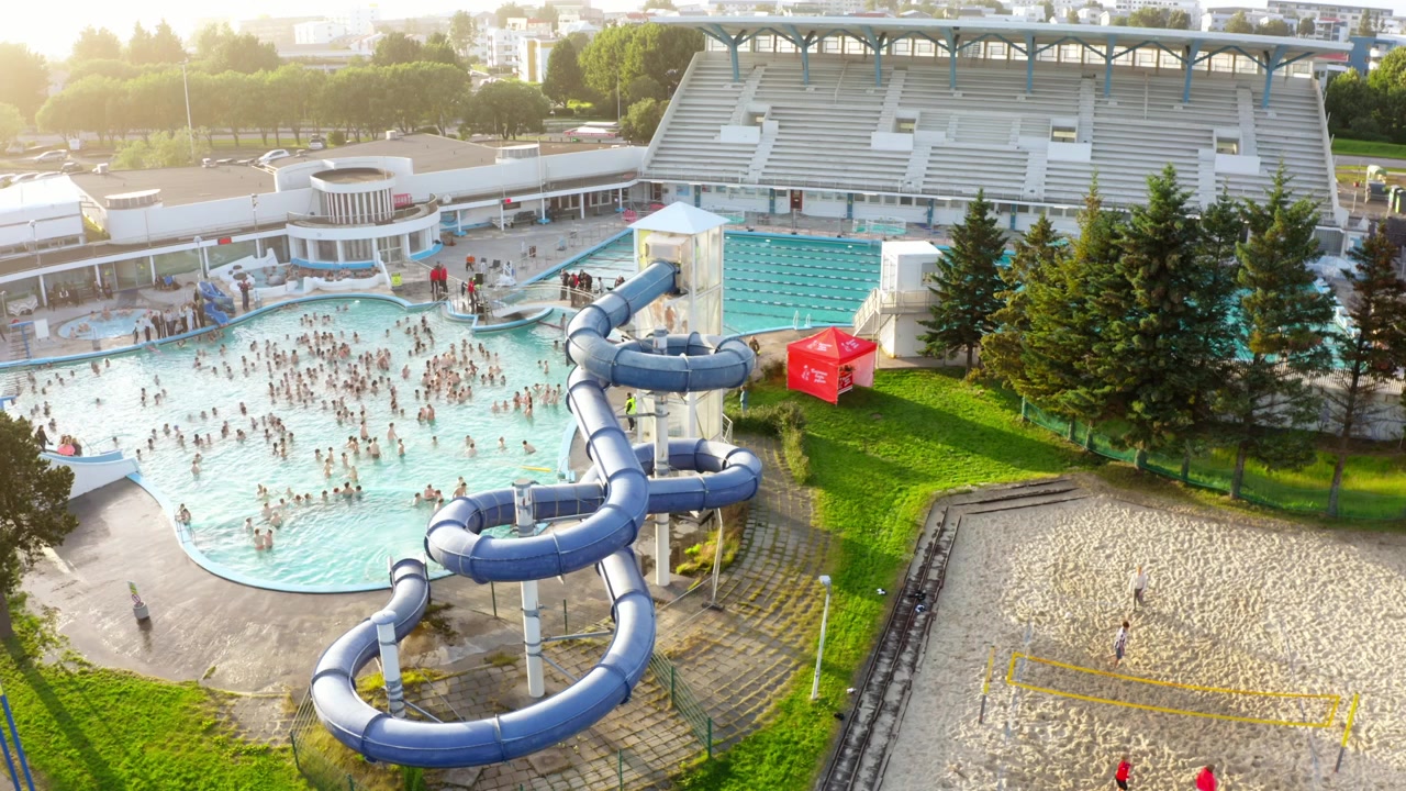 En flygbild visar en fullsatt offentlig simbassäng i Reykjavik, Island. Människor simmar, använder en vattenrutschkana och spelar volleyboll bredvid en stor stadion en solig dag