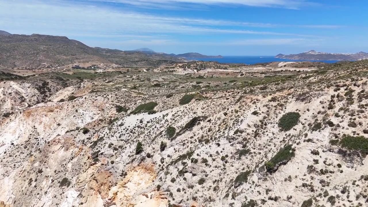Riprese aeree sopra Paralia Firiplaka a Milos, Grecia, rivelano calanchi erosi e pallidi, arbusti sparsi, un terreno vulcanico ondulato e una striscia di Mar Egeo blu intenso in lontananza.
