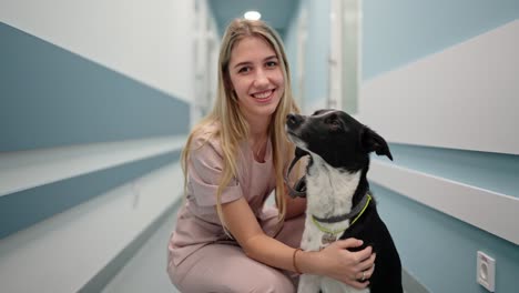 Portrait-of-a-happy-blonde-veterinarian-girl-in-a-pink-uniform-with-a-black-and-white-dog-in-the-corridor-of-a-veterinary-clinic