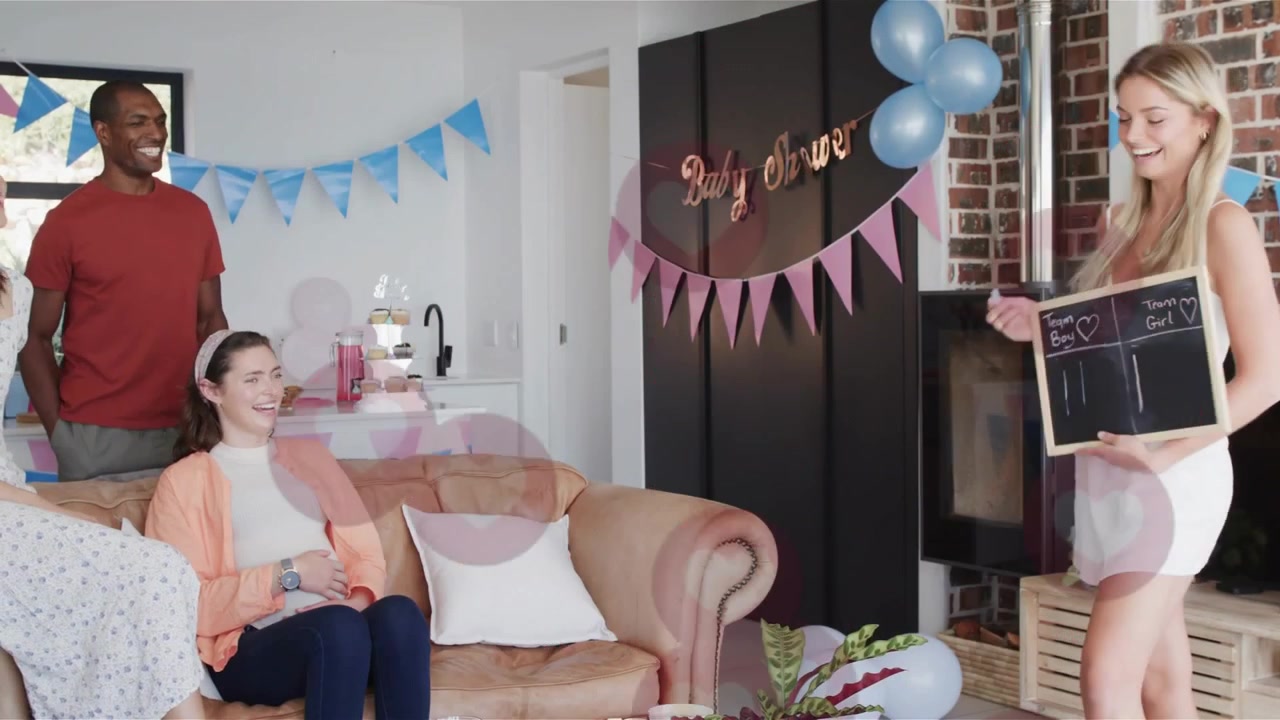 Video vertical: Anfitriona con un cartel, vestida de blanco, dirigiendo un juego de baby shower en casa, con corazones flotando