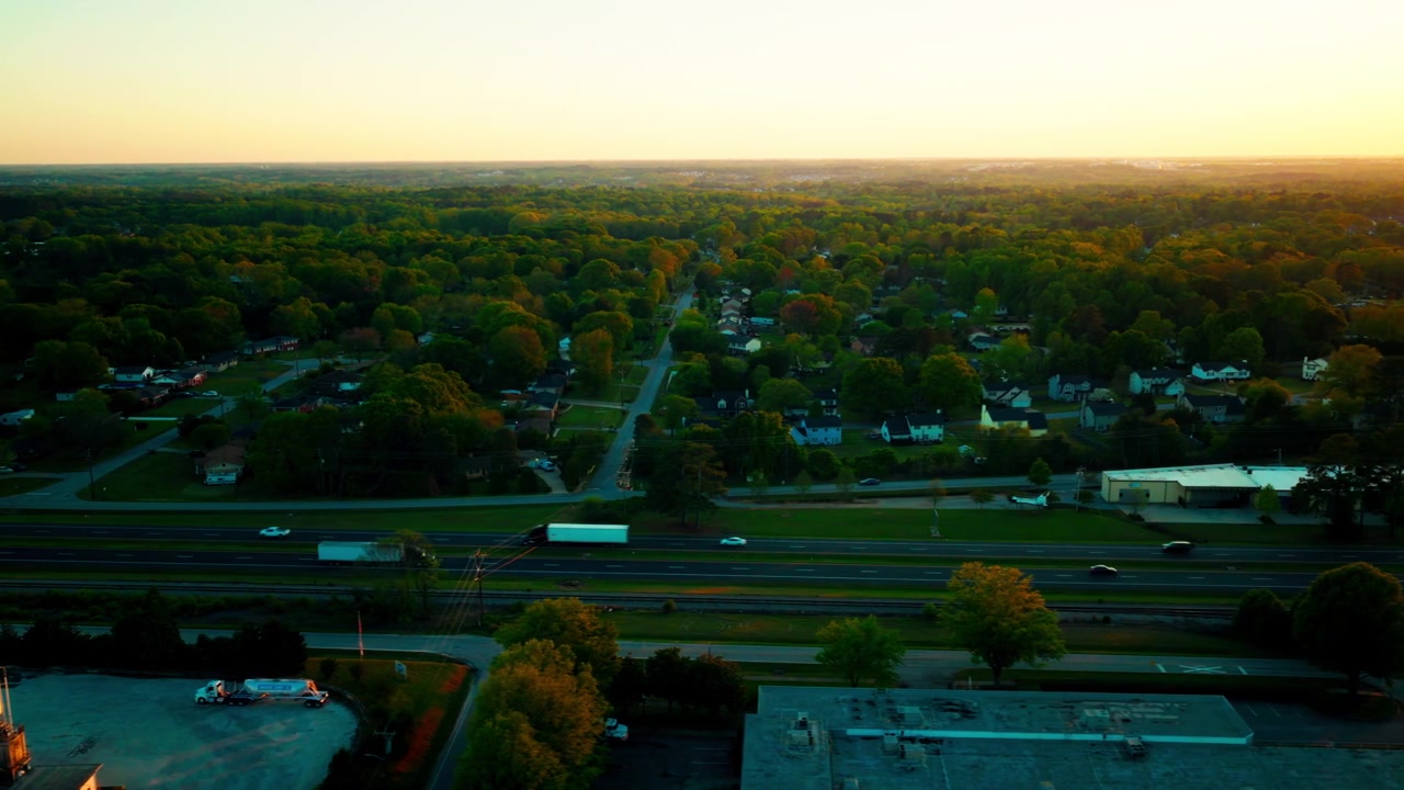 Ampia ripresa aerea cinematografica all'ora d'oro su Greenville, Carolina del Sud, che segue un camionista, autostrada divisa con traffico leggero, binario ferroviario parallelo, quartiere residenziale consolidato e vasto orizzonte boschivo.