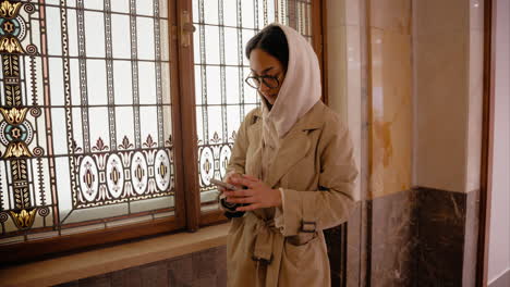 ein junges mädchen scrollt auf ihrem handy und steht neben einem fenster mit einem kapuzen und einem beigenen trenchcoat