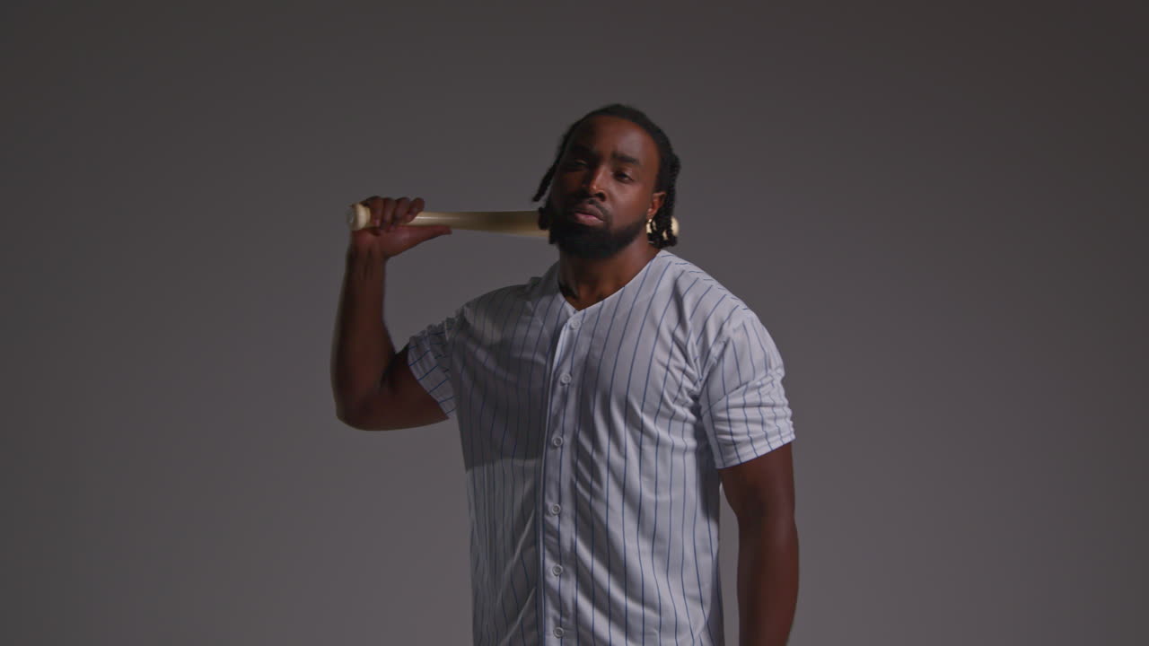 retrato de estudio de un jugador de béisbol masculino serio con una camiseta del equipo sosteniendo un bate girando disparado contra un fondo gris