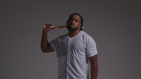 Studio-Portrait-Of-Serious-Male-Baseball-Player-Wearing-Team-Shirt-Holding-Bat-Turning-Around-Shot-Against-Grey-Background