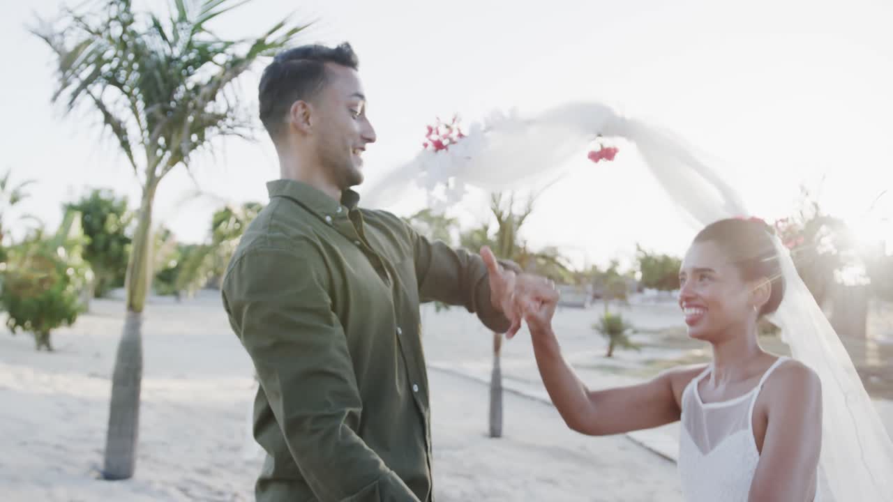 Happy diverse bride and groom dancing and smiling at their beach wedding, in slow motion