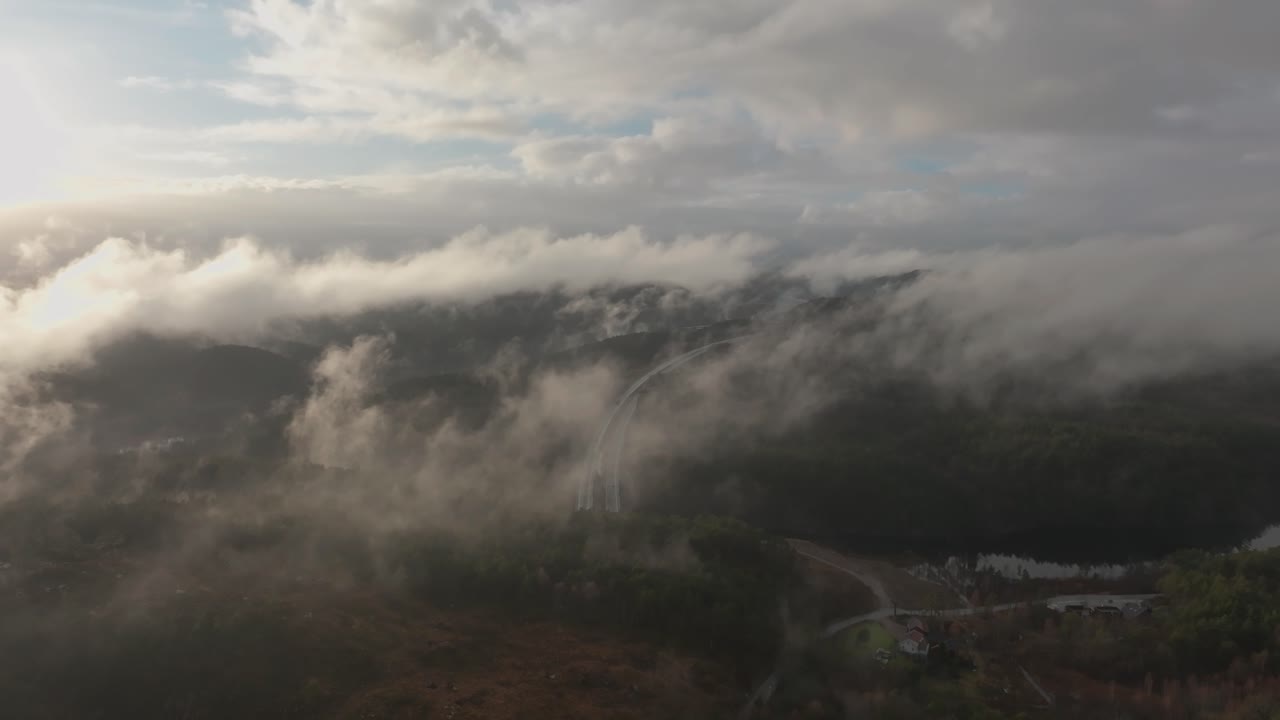 imágenes de drones de la carretera y la urbanización en noruega con algo de niebla flotando en el aire y el sol iluminando las nubes