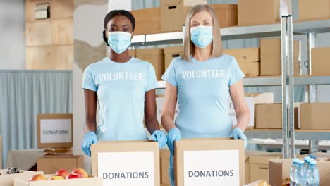 voluntarias afroamericanas y caucásicas con máscaras faciales empacando cajas de donación y mirando la cámara en un almacén de caridad