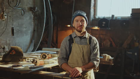 portrait of young man in apron posing at camera in blacksmith workshop