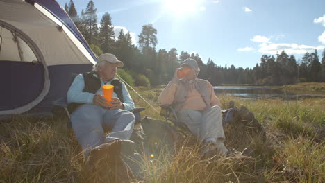 Senior-couple-sitting-outside-a-tent-drinking-near-a-lake