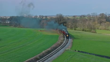 una vista aérea de un tren de pasajeros de vapor que viaja soplando humo, en una sola vía, llegando alrededor de una curva en un soleado día de primavera