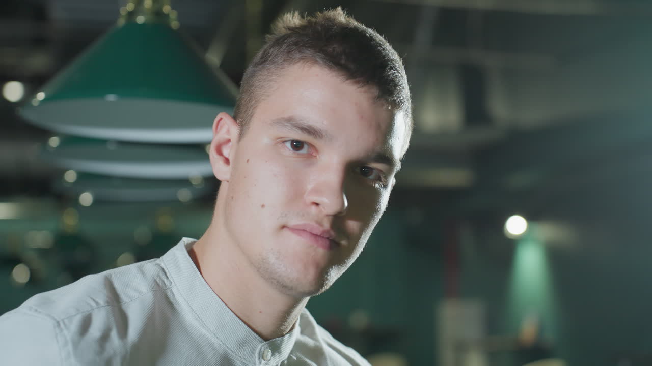 색 셔츠를 입고 약간 미소 짓는 남자의 클로즈업, 희미하게 밝은 빌리어드 홀, 오버헤드 조명은 그의 얼굴에 부드러운 반사를 만니다. 초록색 빌리드 테이블과 매달린 조명은 현대적인 분위기에 깊이를 더합니다.