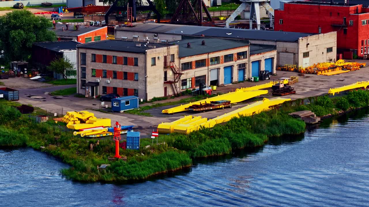 An aerial shot shows a section of the industrial Port of Riga in Latvia, with workshops, warehouses, and large, unassembled crane parts stored on the riverbank