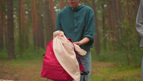 hikers walk through a serene forest, carrying backpacks, one in a green shirt and the other in a gray sweater , they pause near a fallen tree as the lady in green starts to open her red bag