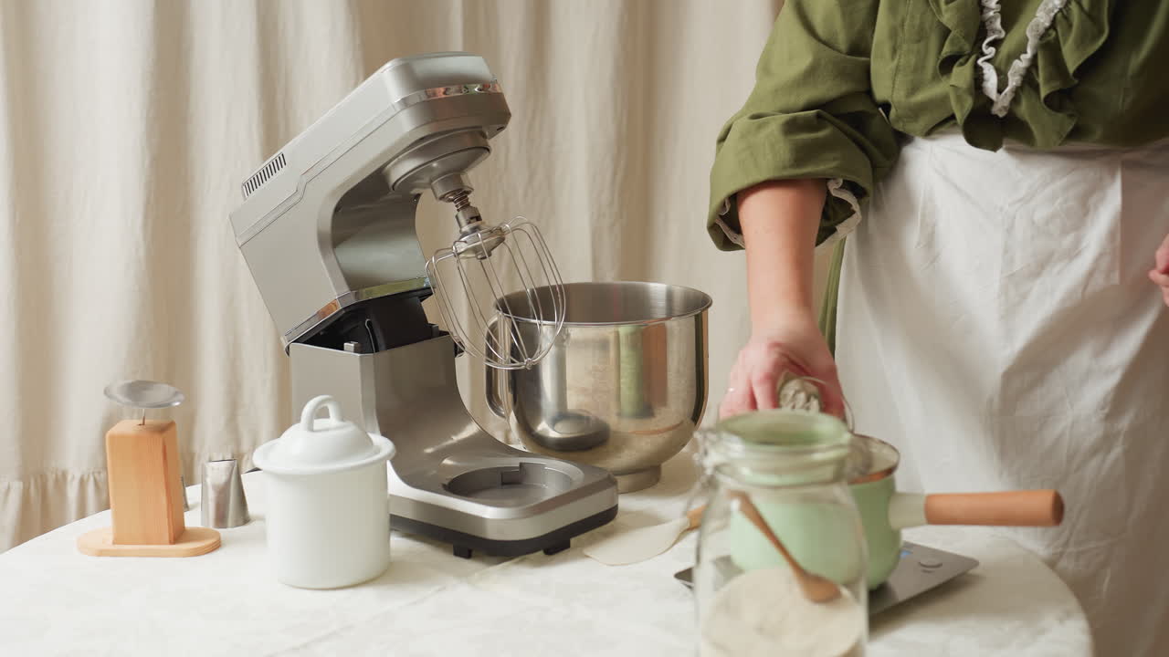Cook in green dress picks up clear glass filled with water and pours it into metal pot on kitchen table beside mixer, jars, and utensils while preparing ingredients