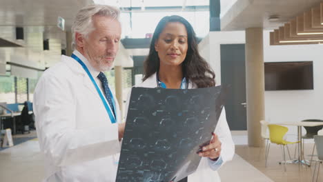 Doctor-Wearing-White-Coat-Discussing-Patient-Scan-With-Female-Colleague-In-Busy-Hospital