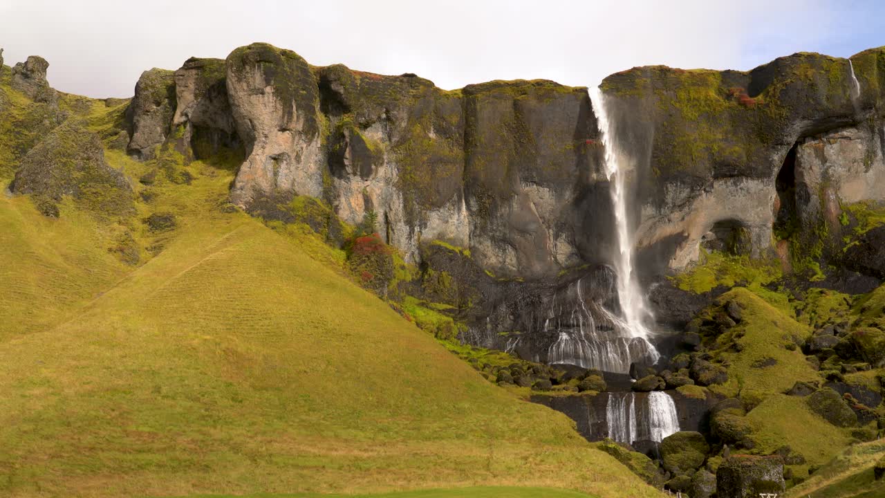 Acqua di una cascata islandese soffiata dal vento mentre cade da una scogliera a picco, in alto sopra un verde pascolo d'erba e cascate a gradoni più piccole sottostanti.