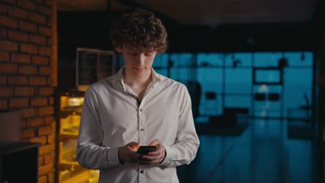 Confident-guy-with-curly-hair-in-a-white-shirt-typing-on-his-phone-and-standing-in-the-evening-office