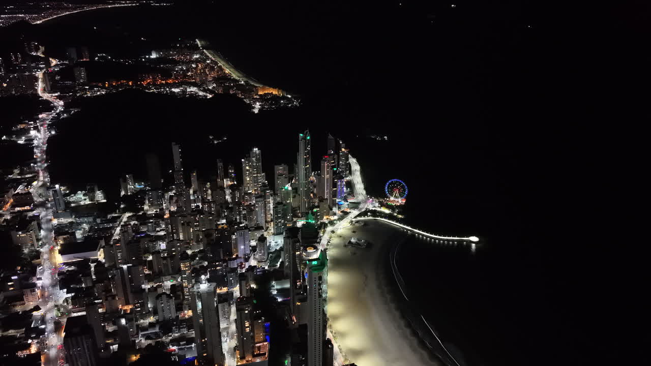 Nighttime drone pan reveals glowing skyline, Ferris wheel, and brightly lit Avenida Atlântica beachfront of Balneário Camboriú, Santa Catarina, Brazil, with the coastline curving into the distance