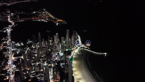 nighttime drone pan reveals glowing skyline, ferris wheel, and brightly lit avenida atlântica beachfront of balneário camboriú, santa catarina, brazil, with the coastline curving into the distance
