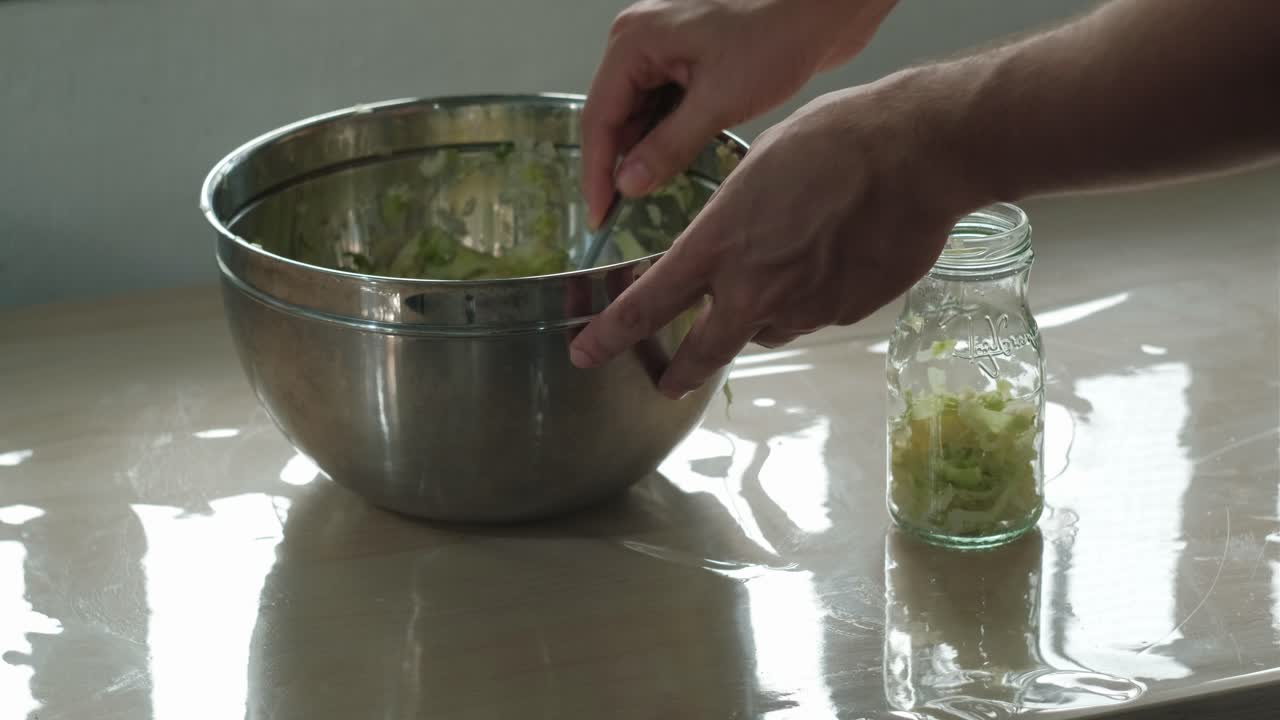 Hands and spoon scooping prepared sauerkraut from bowl into glass mason jar for storage - static medium shot