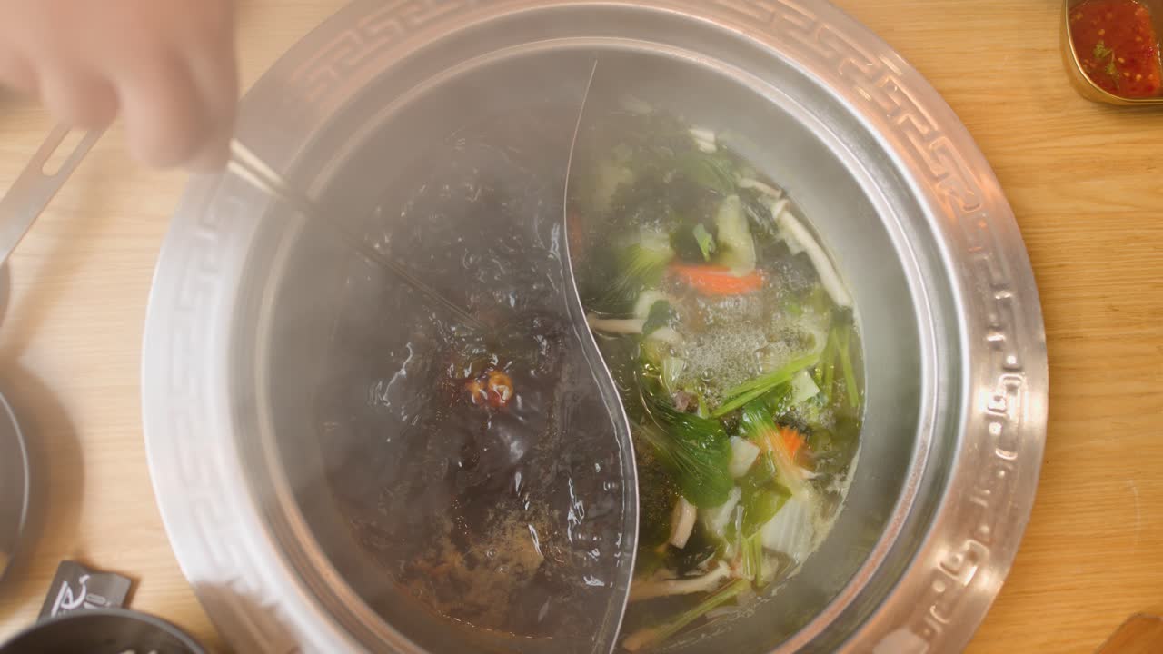 Hand cooks sliced beef in steaming dual broth hotpot, overhead view, warm ambient lighting