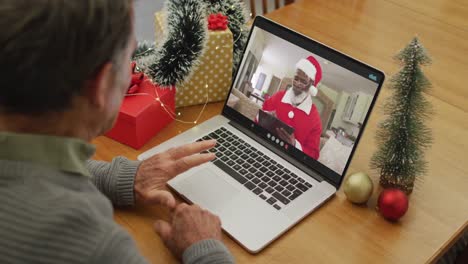 Un-Hombre-Mayor-Caucásico-Teniendo-Una-Videollamada-Navideña-En-Una-Computadora-Portátil-Con-Un-Papá-Noel-Americano-Africano-En-La-Pantalla