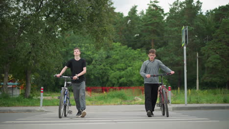 two brothers walking side by side with their bicycles across a pedestrian crossing marked with white paint, one brother in a grey top is seen climbing onto his bike, while a car passes by