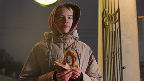 elegant woman standing outdoors with pastry in hand, looking content, dressed warmly, smiling in front of an illuminated clock. urban setting at night with cozy ambiance