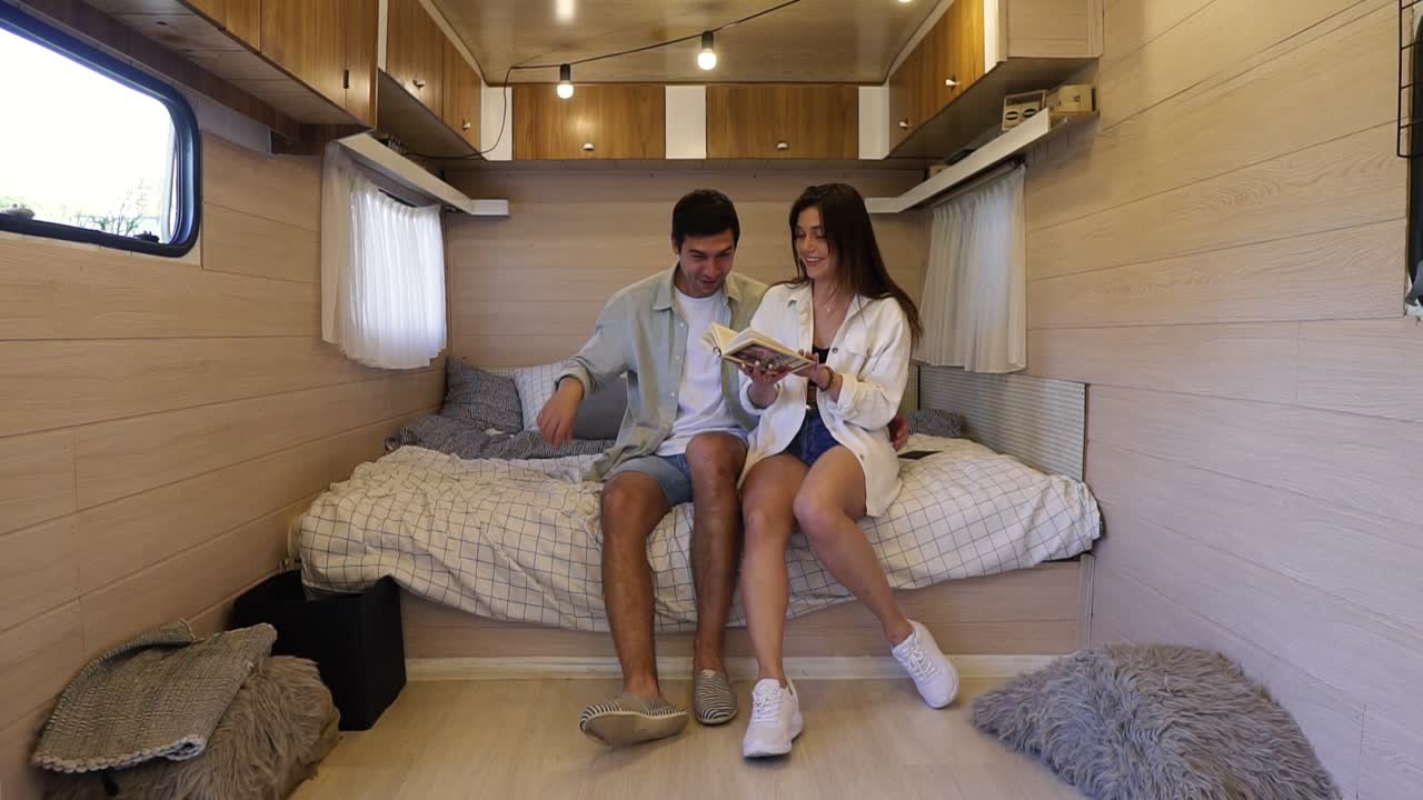 Cheerful, european couple sitting on the big bed inside the wheels house, having time together - reading book, having fun. Recreation, vacation, travelling in van