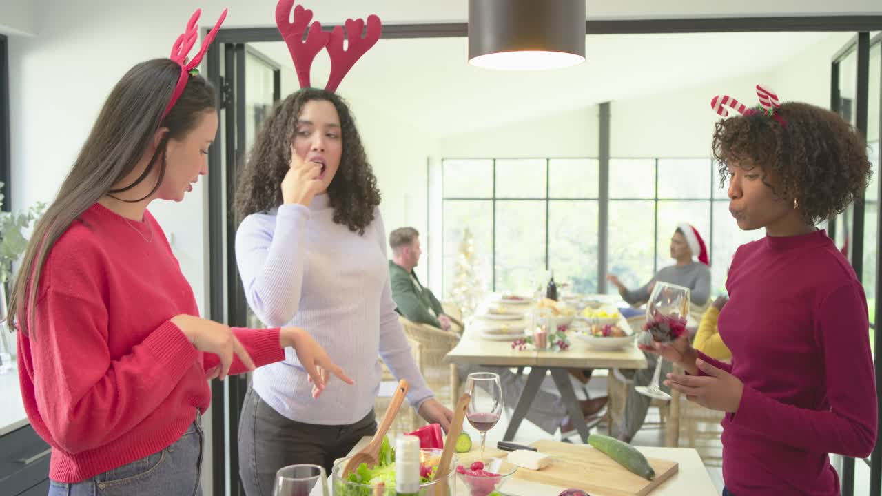 Female friends donning antlers toasting wine, chopping and tossing salad at kitchen island