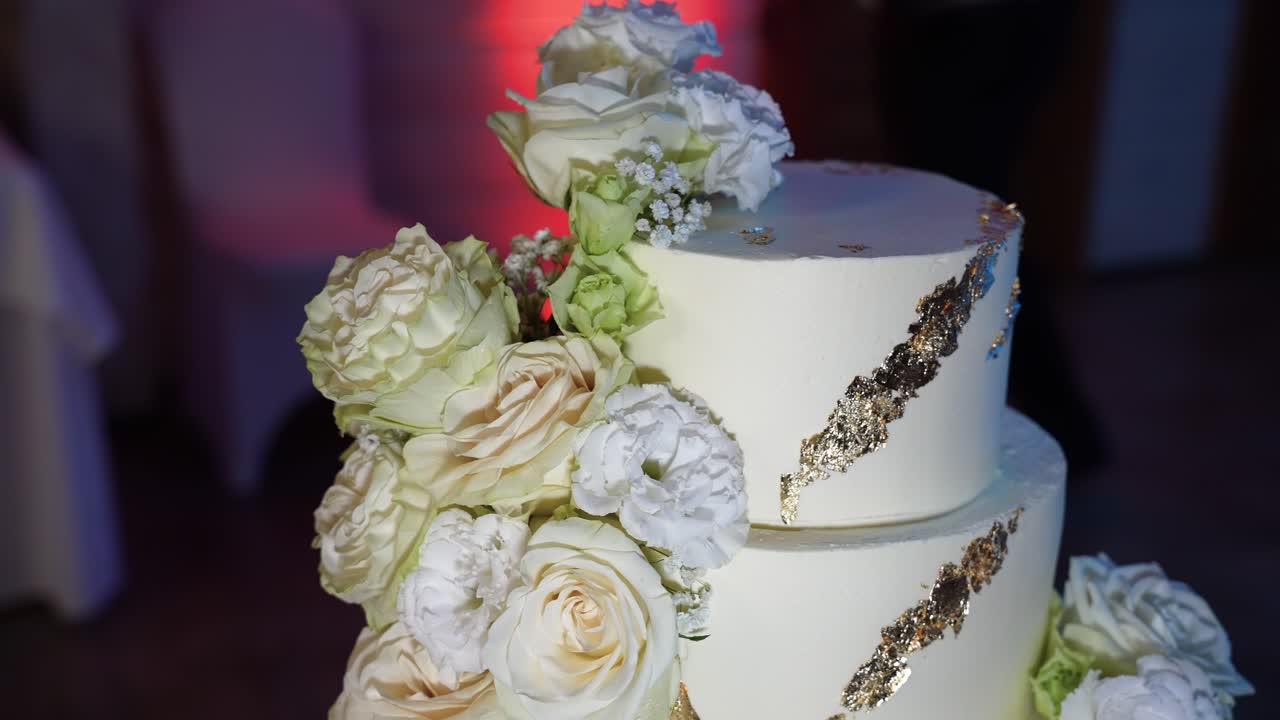 Close-up de um bolo de casamento lindamente elaborado com delicadas rosas brancas e detalhes dourados ornamentados. O design luxuoso é realçado por uma iluminação suave, criando um ambiente romântico
