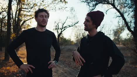 a brunette guy in a red hat talks to his friend with a beard in a black sports uniform who tells him how to properly go for a morning jog in the fall during sunrise