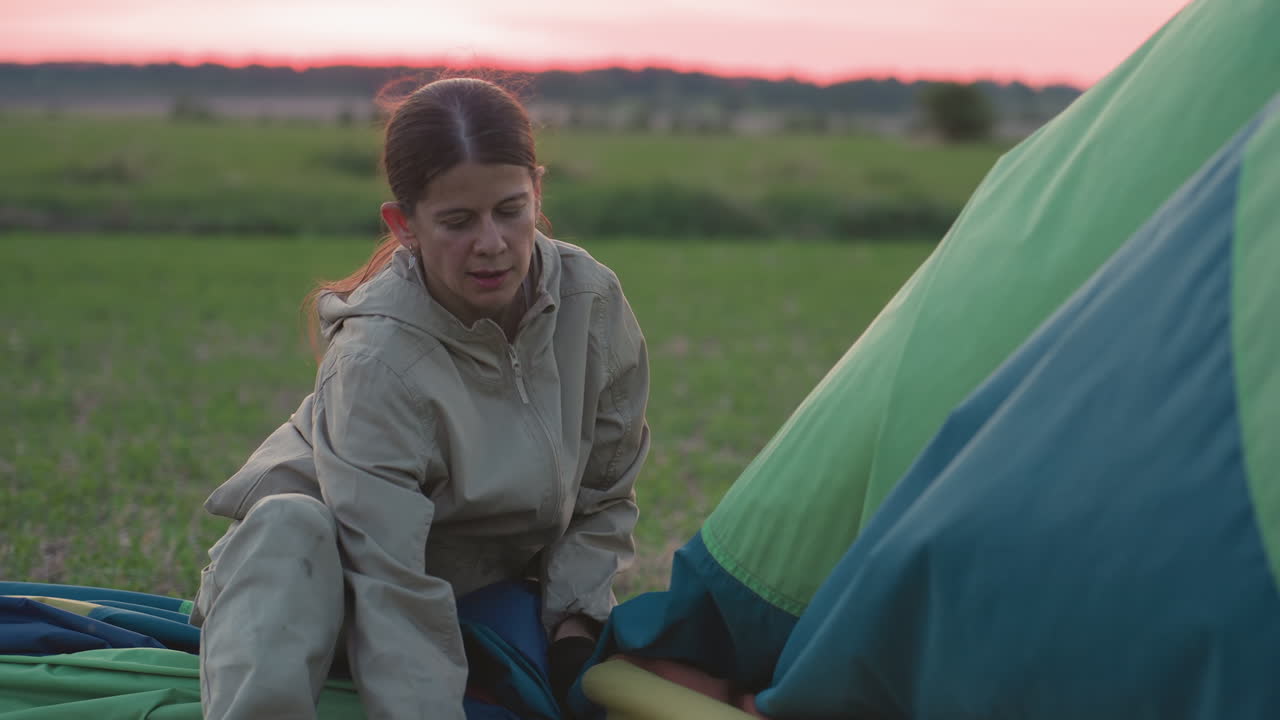 woman crouching on expansive green field packing deflated multicolour hot air balloon canopy into compact folds along sturdy ropes under soft pink dusk sky