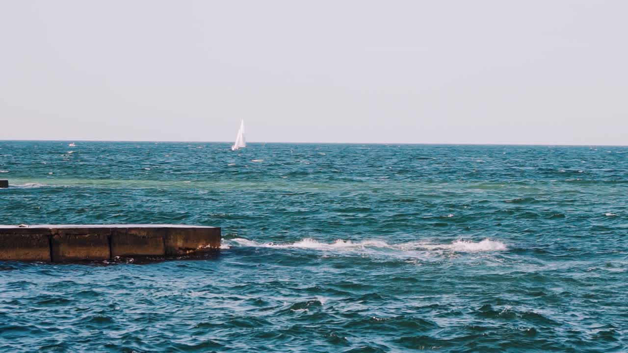 Motion of sea water. Waves crashing on stone harbor making white foam. White sail floating on the water surface. Slow motion.