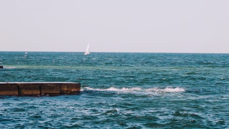 motion of sea water. waves crashing on stone harbor making white foam. white sail floating on the water surface. slow motion.