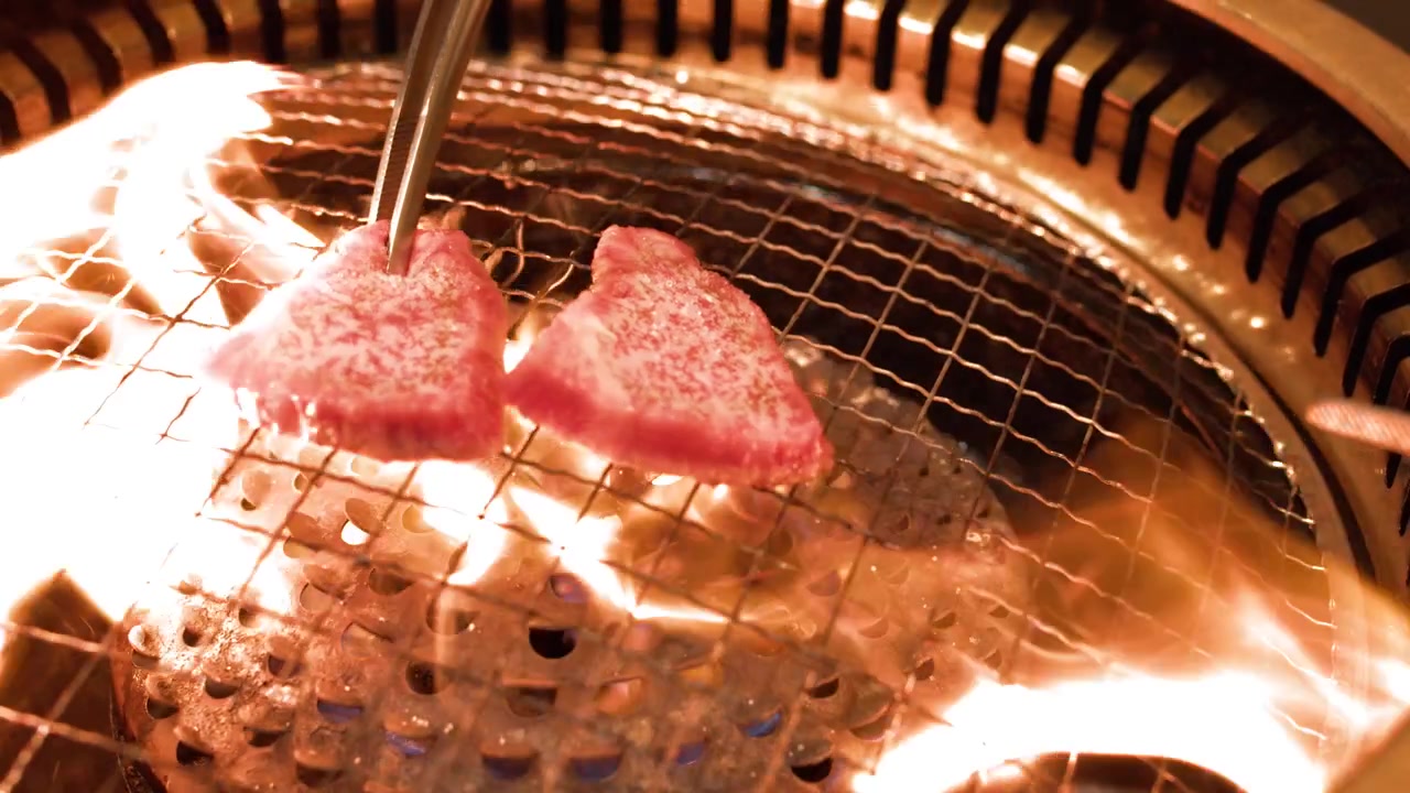 激しいオレンジ色の炎と煙が立ち上る丸い金属製グリルで焼かれる霜降り牛肉のステーキ。