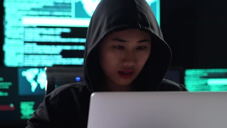 close up of asian female hacker in a hood celebrating succeed attack global infrastructure servers by a laptop with maps and data on display screens of multiple monitors in a dark office room