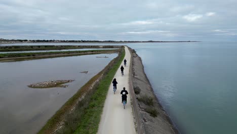 Familia-Montando-En-Bicicleta-En-El-Pueblo-De-Loix-En-La-Isla-De-Île-De-Ré-En-El-Oeste-De-Francia-Mientras-Un-Niño-Pequeño-Sostiene-La-Mano-De-La-Madre,-Seguimiento-Aéreo-Detrás-De-La-Toma