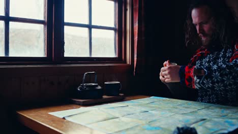 man in nordic sweater pouring coffee and milk into mug on wooden table by the window while looking at map spread out in front. medium shot