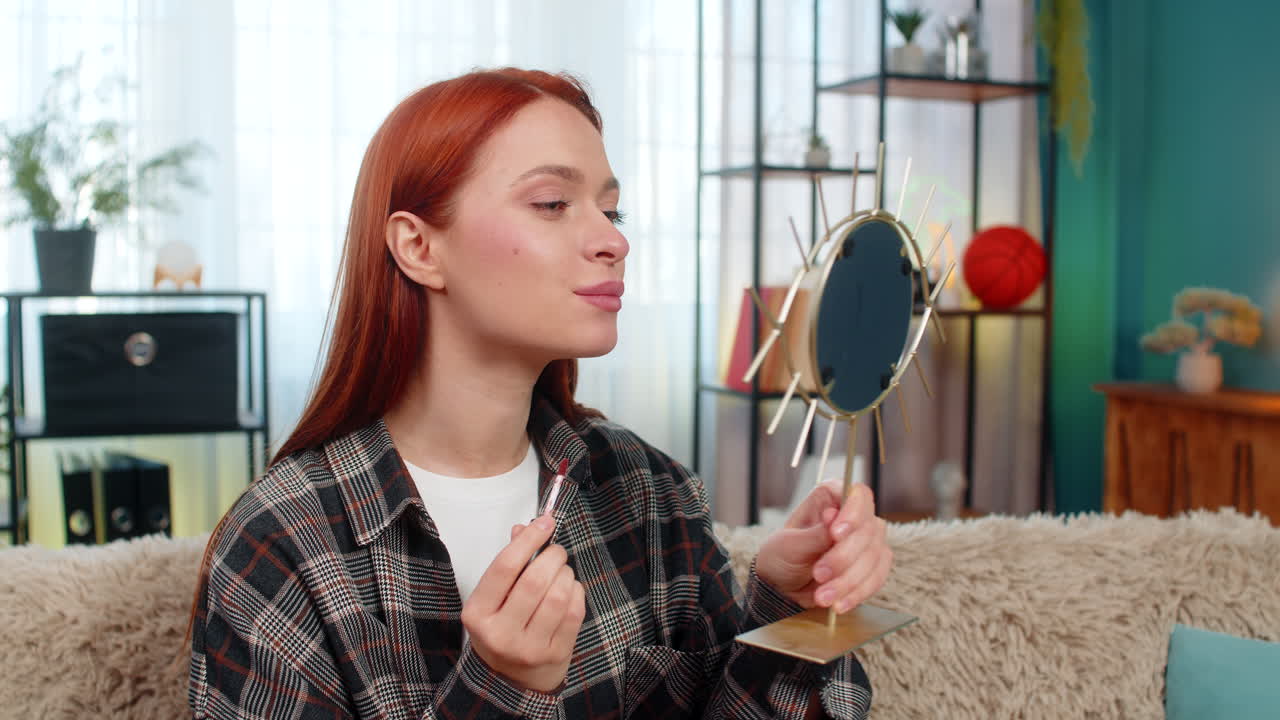 Young pretty woman applying makeup looking in mirror smiling while drawing lips with lipstick gently