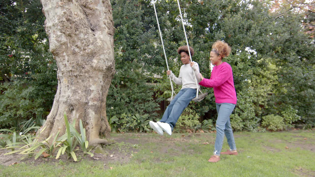 Madre afroamericana felice che spinge il figlio sull'altalena dell'albero in giardino, al rallentatore.