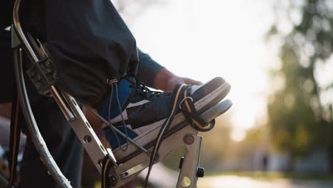 primer plano de la mano masculina ajustando las correas de los zancos con zapatillas de deporte negras y azules visibles. lleva una sudadera gris con capucha, con la luz del sol iluminando suavemente la escena y la vegetación borrosa en el fondo