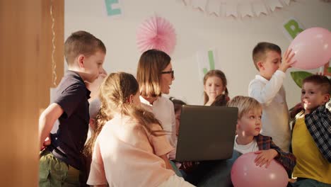 una maestra con un peinado bob y gafas sostiene una computadora portátil gris delante de ella y se comunica con los niños en la habitación para prepararse para la escuela en un club para niños en edad preescolar. lección en el club para preparar a los niños para la escuela