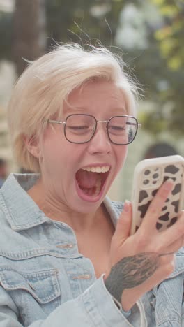 feliz mujer rubia caucásica usando teléfono inteligente celebrando ganar buenas noticias mensaje en la ciudad calle de la ciudad