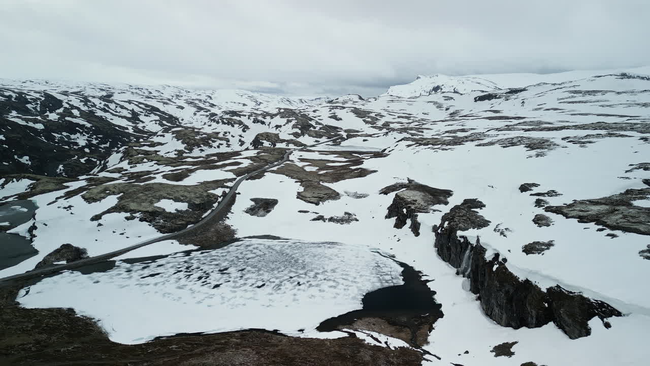 Drone shot of the Aurlandsfjellet tourist road in Norway. The snow-covered landscape is vast. In the foreground is an ice-covered lake with a waterfall