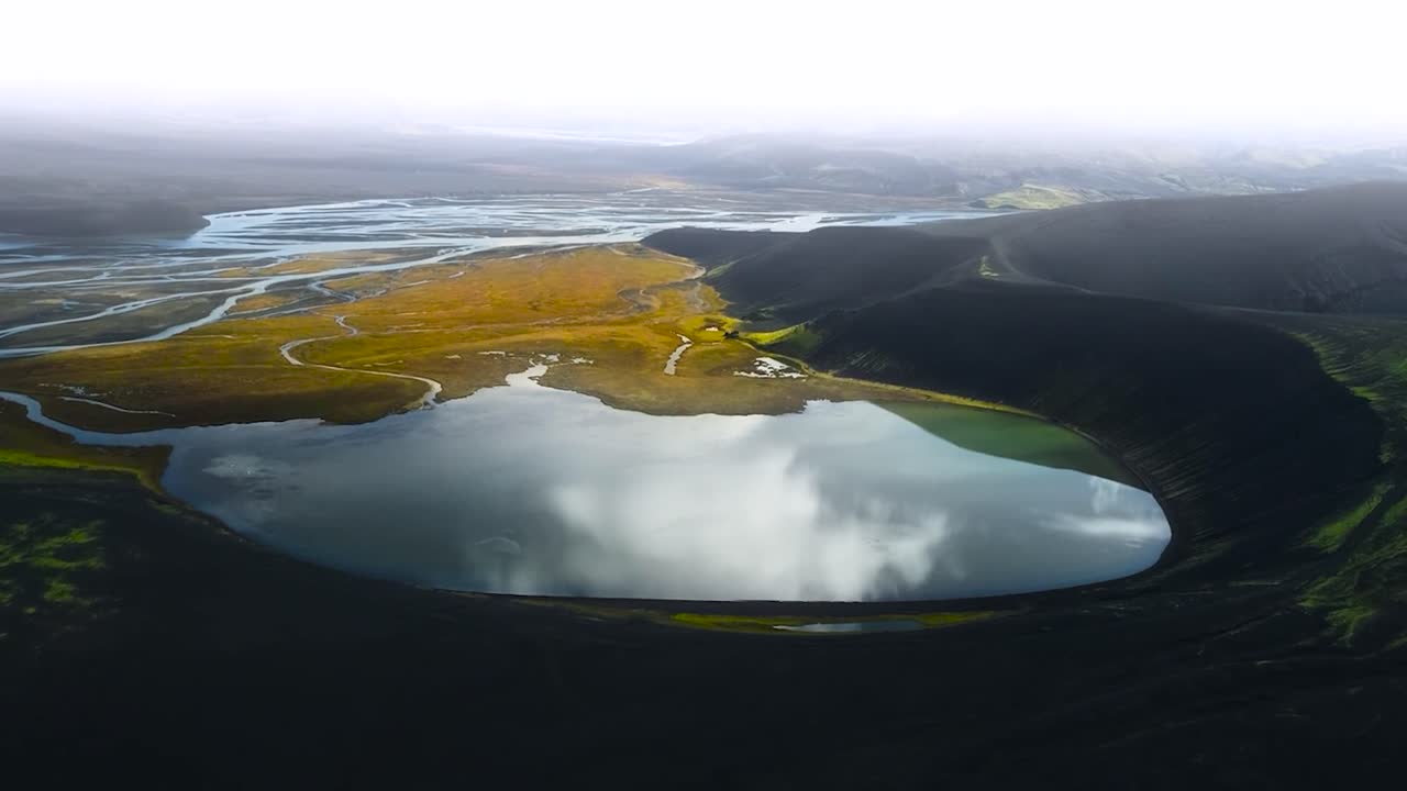Aerial drone footage of a large lake in a crater that has sharp steep mountain cliff edges covered in green moss and plants. River has brown plants and grass around in Greenland or Iceland, cloudy day
