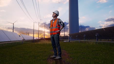 full body side view of asian male engineer wearing safety helmet looking around while standing with arms akimbo with solar panel and wind turbines