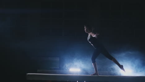 la niña realiza un truco en un tronco con luz de fondo y cámara lenta en ropa de gimnasia deportiva. humo y azul. saltar y girar en la viga de equilibrio