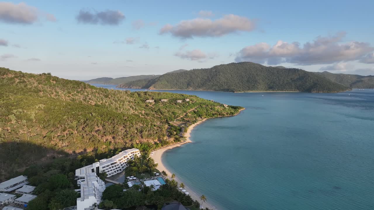Flycam lùi chậm trong khi bay vòng sang phải, hé lộ bờ biển nhiệt đới của Đảo Hayman, bãi biển cát và khu nghỉ dưỡng sang trọng bên cạnh làn nước biển màu ngọc lam tĩnh lặng ở Whitsundays, Queensland, Úc.
