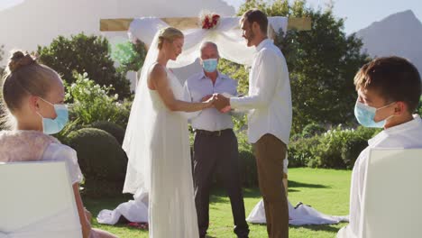 Caucasian-bride-groom-and-wedding-officiant-wearing-face-mask-standing-at-outdoor-altar